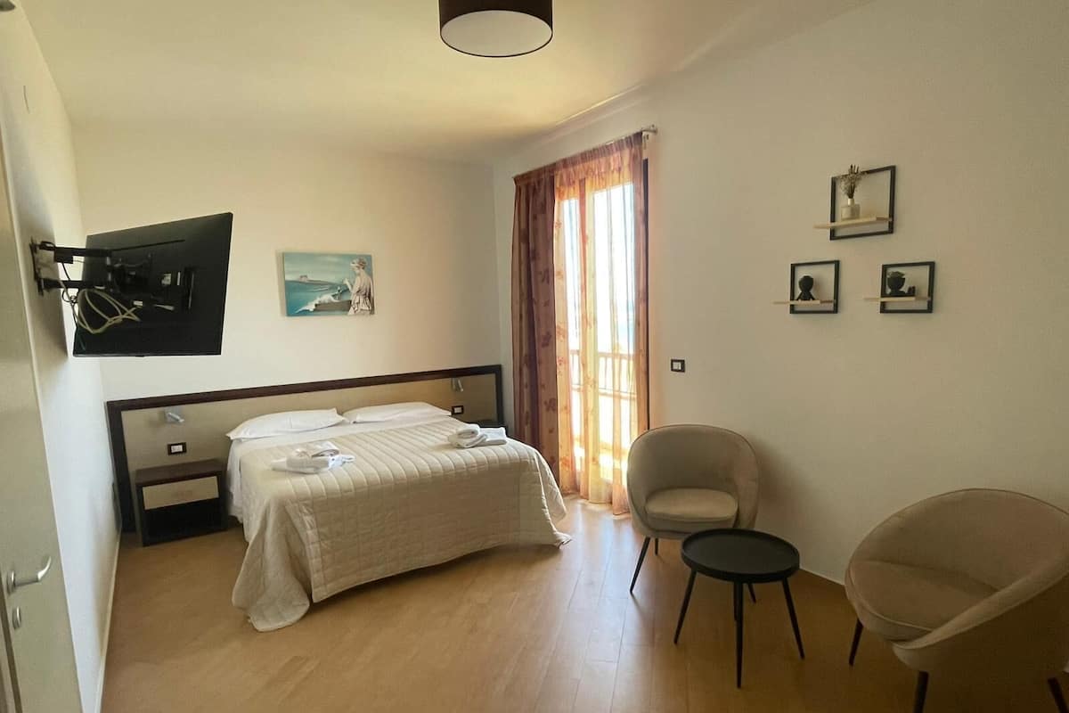 Habitación de hotel con cama, una silla, una mesita, televisión y obras de arte en las paredes.