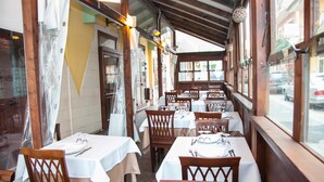 An indoor dining area with tables set for four, wooden chairs, and large windows along the walls.