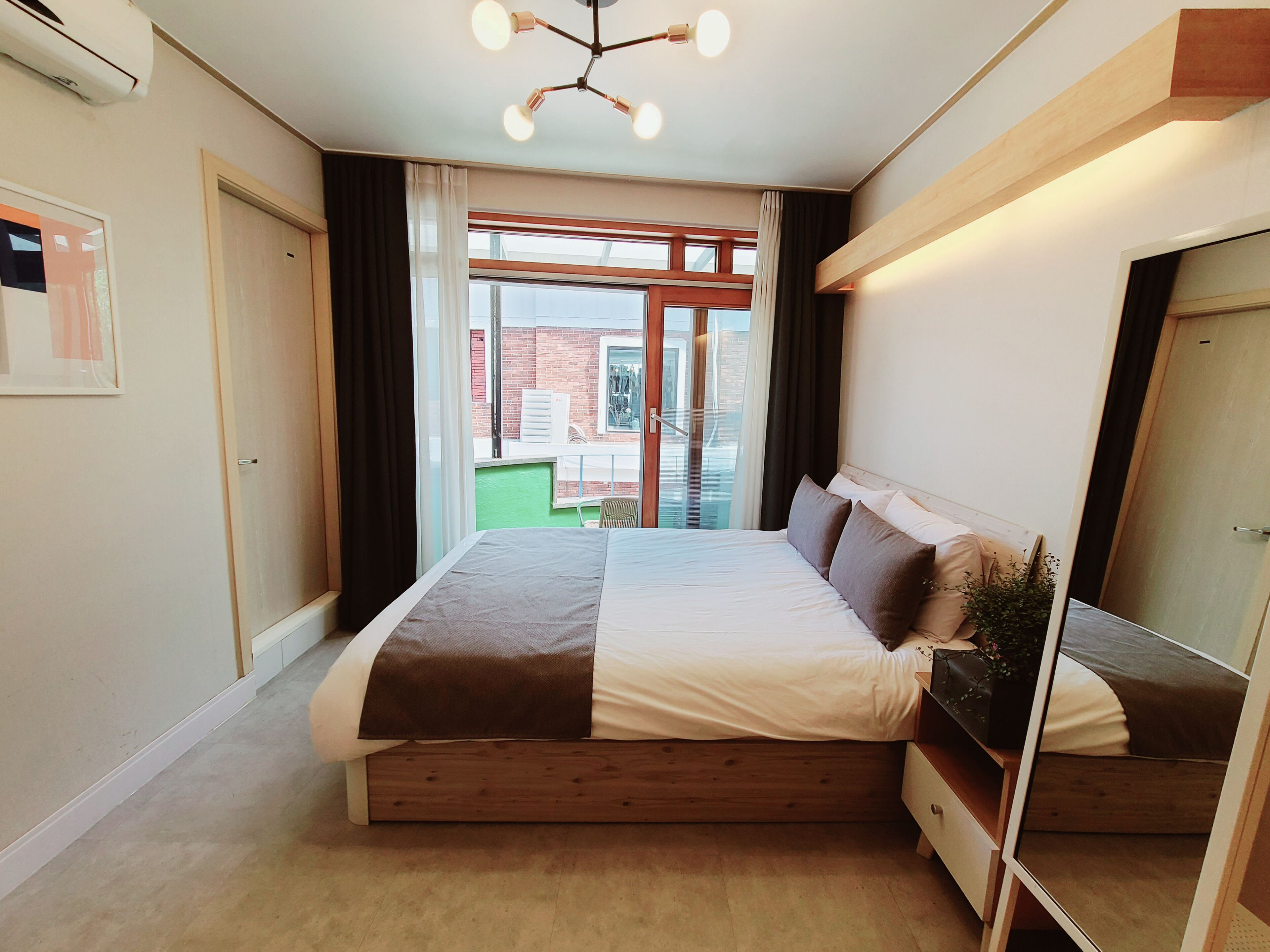 A modern bedroom with a large bed, a wooden headboard, a bedside table, and a view of the outside.