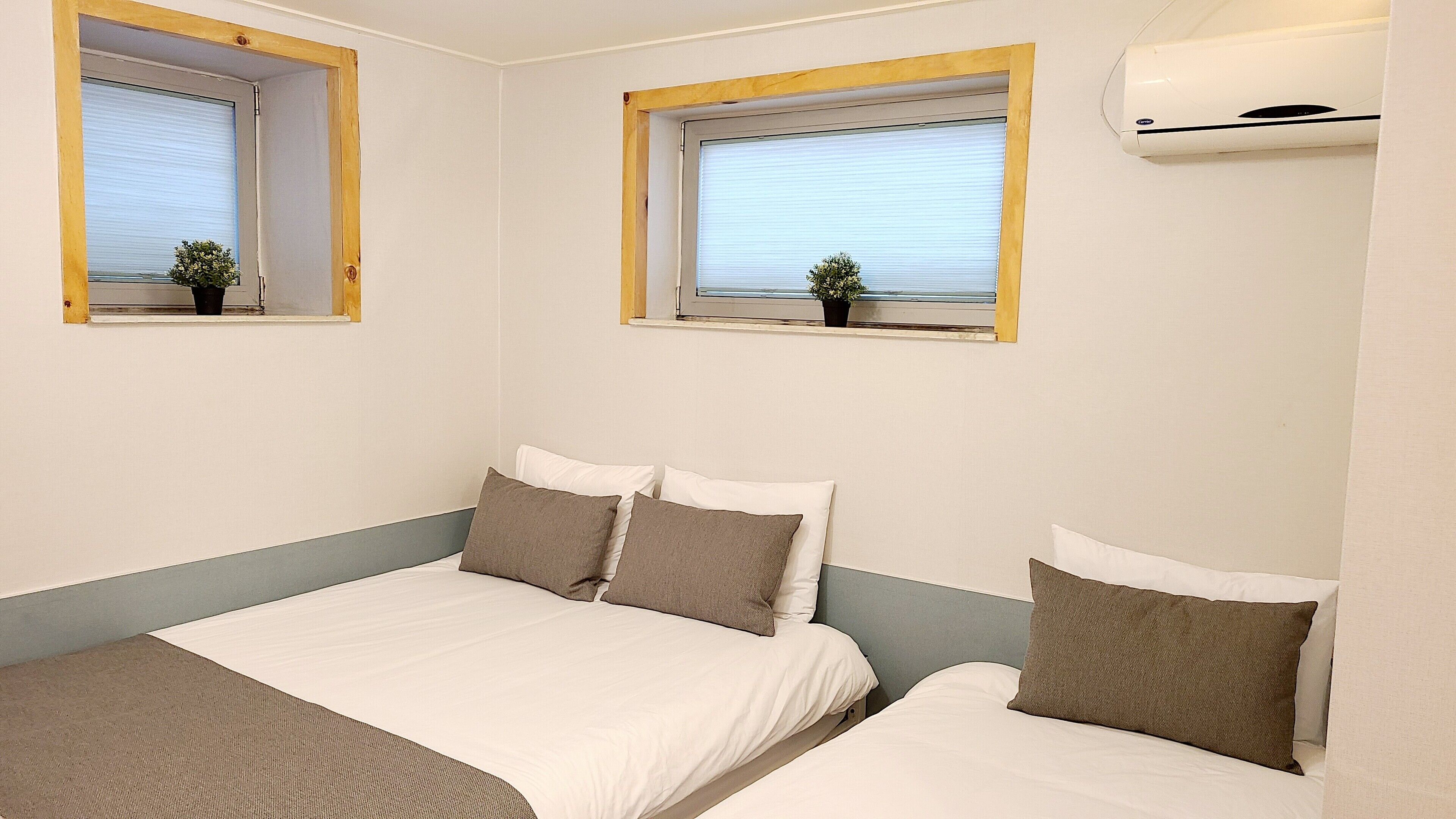 Une chambre avec deux lits, chacun agrémenté de draps blancs et d’oreillers marron. Des fenêtres équipées de stores et de petites plantes en pot sont posées sur les rebords de fenêtre.