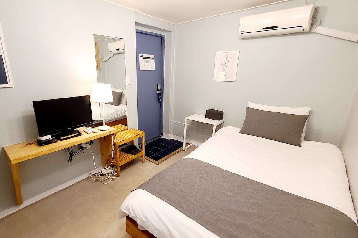 A hotel room with a bed, a desk with a TV, a mirror, an air conditioner, and a door.