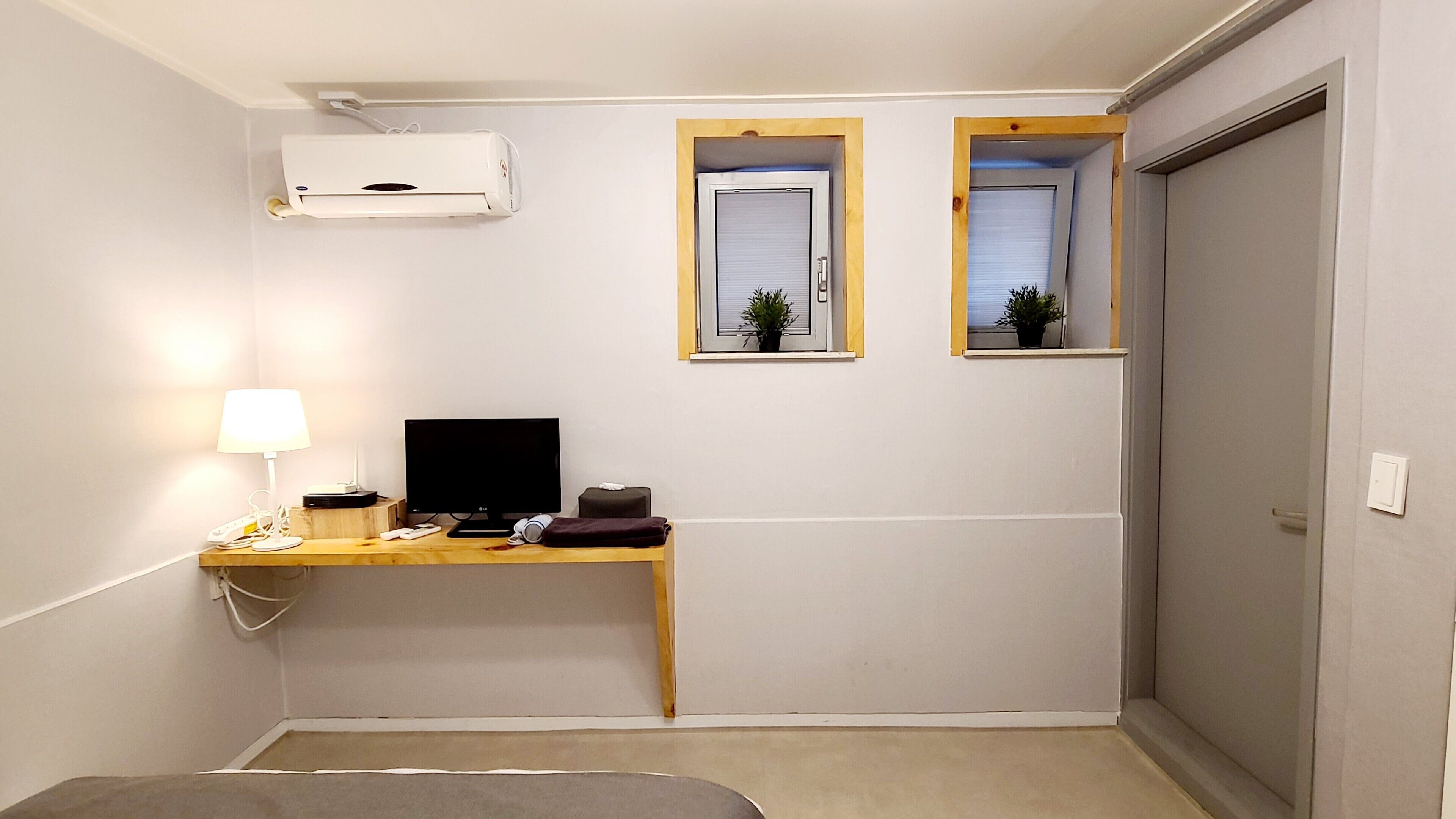 A small, compact room with a desk, a computer, a lamp, and two windows with plants.