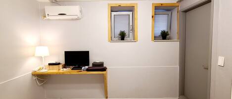 A small, compact room with a desk, a computer, a lamp, and two windows with plants.