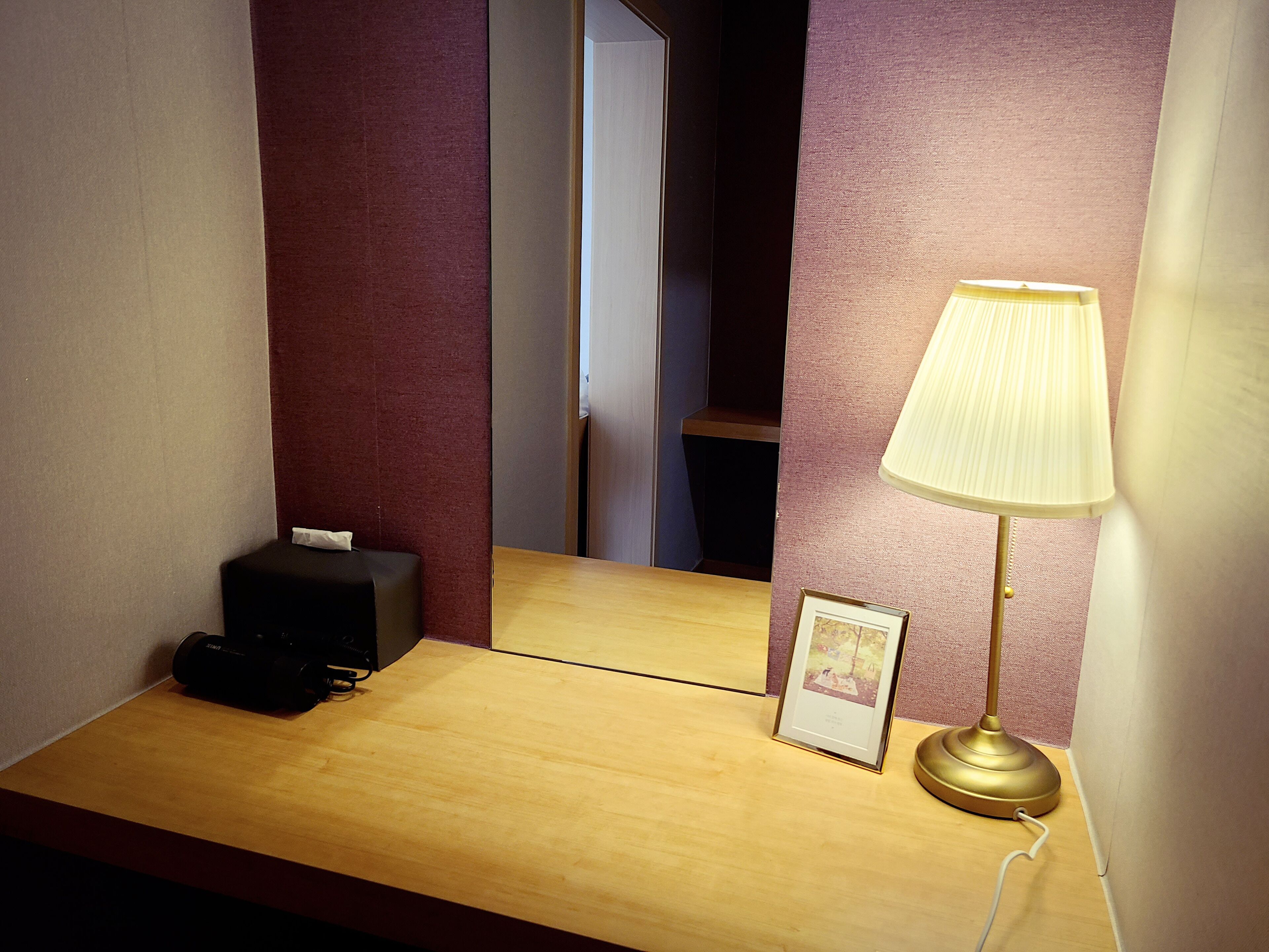 Habitación de hotel con un escritorio de madera, un altavoz negro, un cuadro enmarcado, una lámpara dorada y un espejo que refleja una puerta y una habitación oscura.