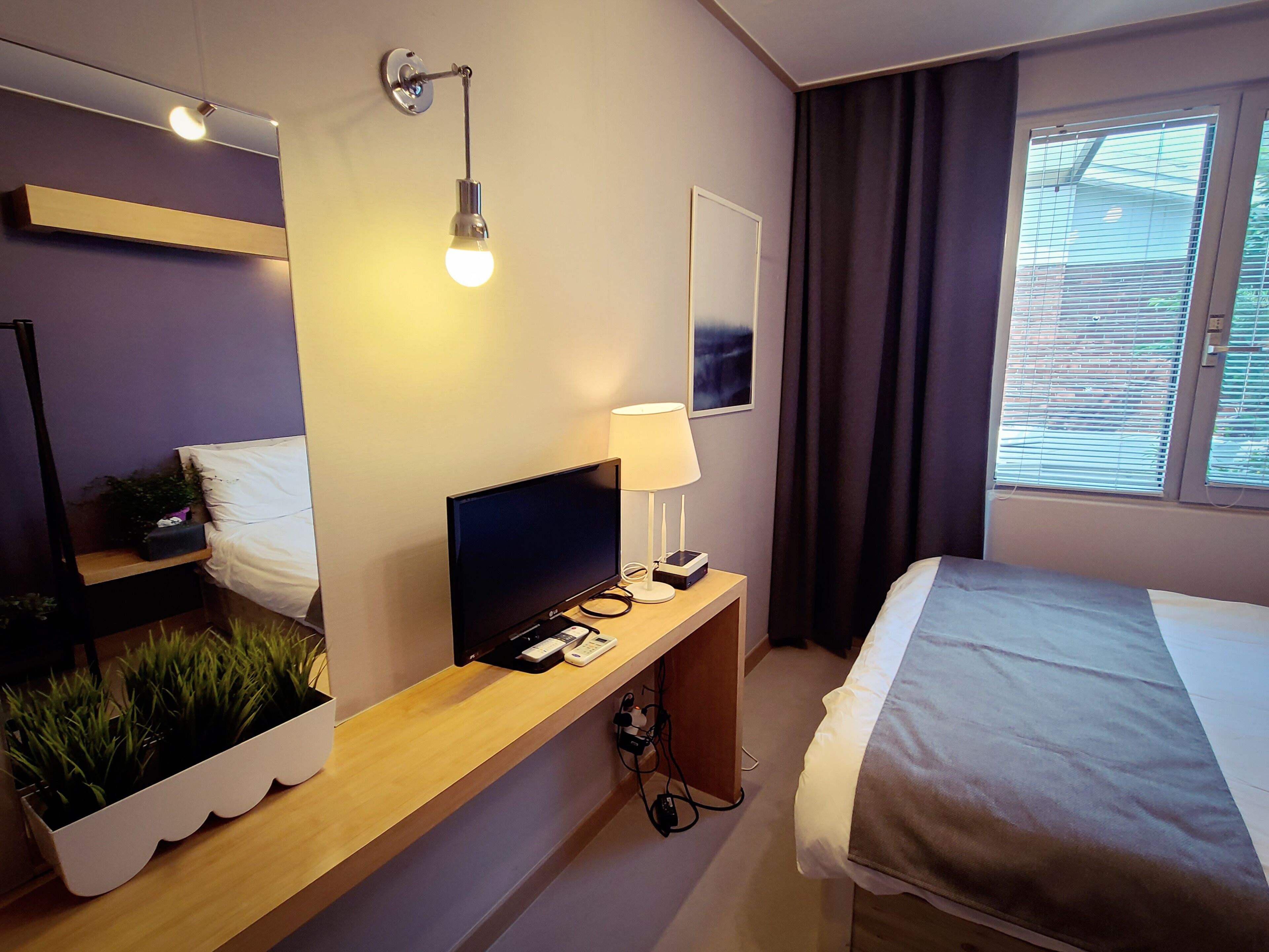 Une chambre d’hôtel comprenant un lit, un bureau avec un ordinateur, une lampe, une télévision, un miroir et une fenêtre avec des rideaux.