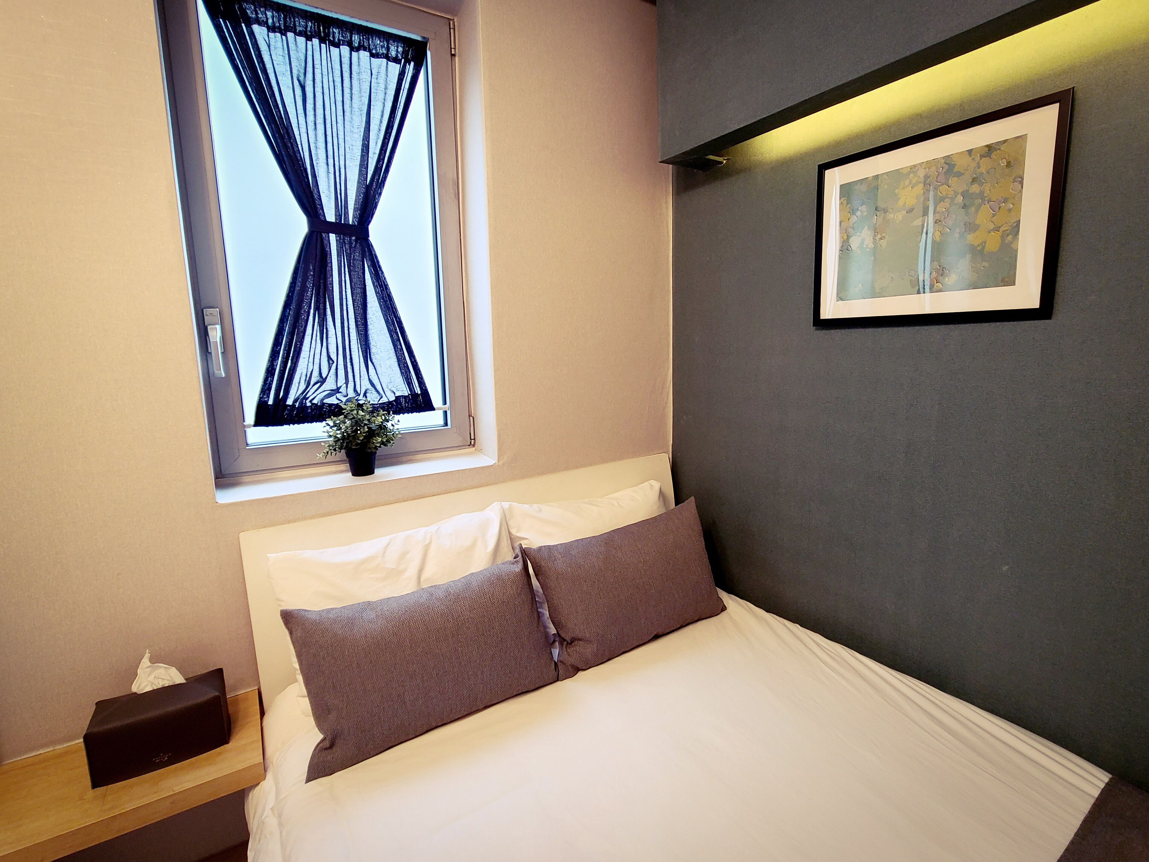 Una cama bien hecha con ropa de cama blanca y marrón, una ventana con cortinas translúcidas, una pequeña planta en maceta y un cuadro en la pared.
