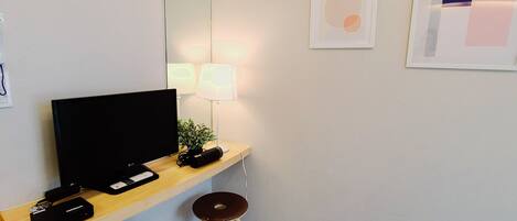 A modern room with a flat-screen TV on a wooden stand, a desk with a lamp, a small stool, and a wall-mounted mirror.