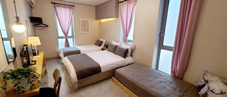 A hotel room with two beds, a desk with a computer, a chair, a plant, and a window with curtains.