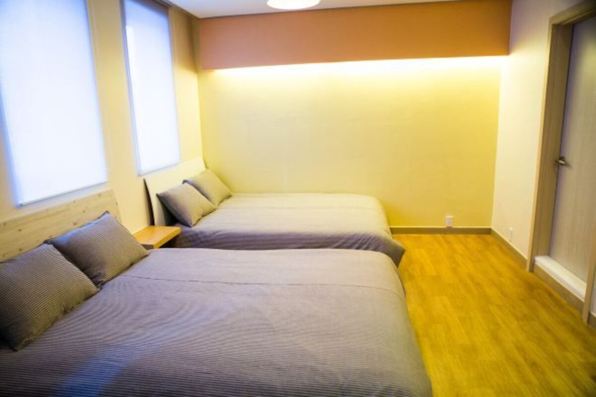 Um quarto pequeno e aconchegante com duas camas, piso de madeira e uma janela com persianas.