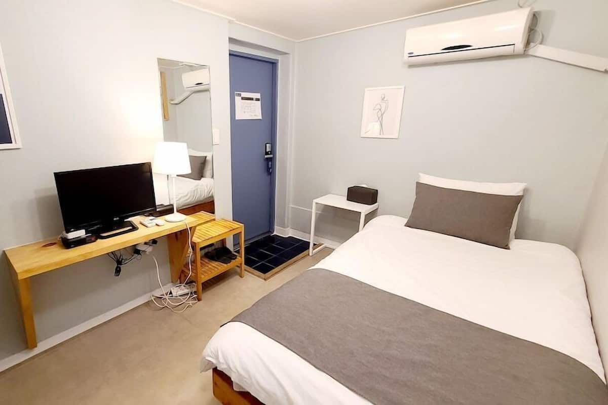 A hotel room with a bed, a desk with a TV, a small table, a chair, an air conditioning unit, and a door.