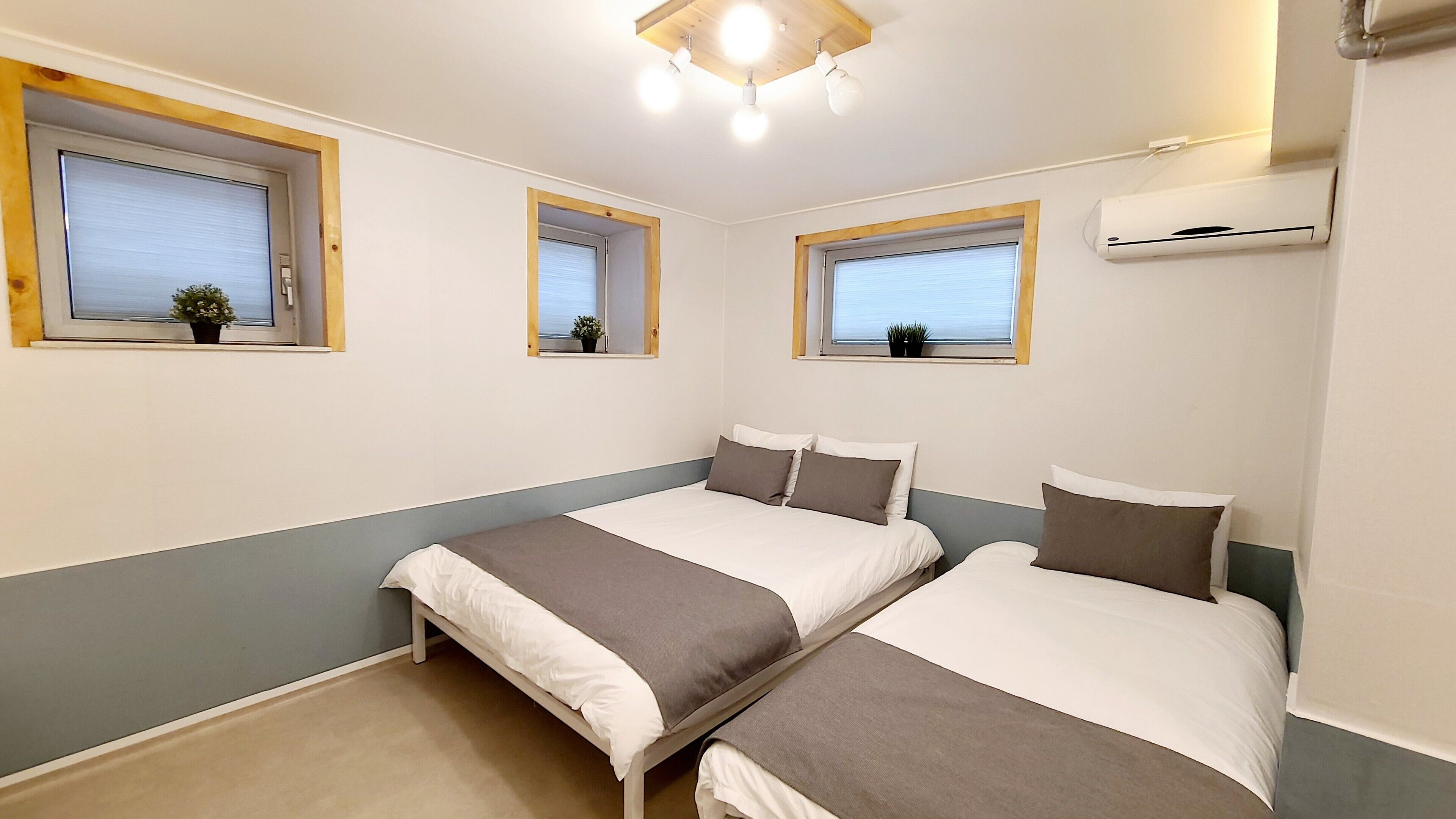A room with two beds, a ceiling light fixture, and three windows with wooden frames.