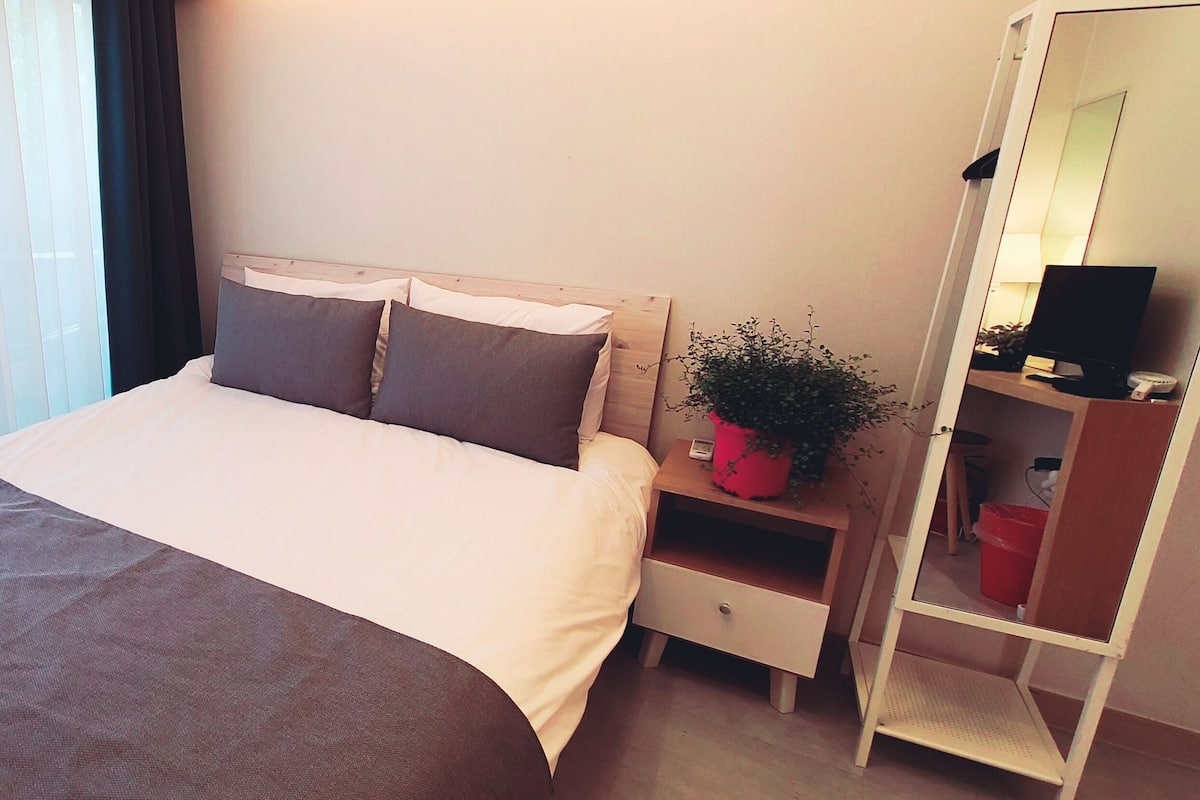 A hotel room with a large bed, a bedside table with a lamp, a small plant, and a wardrobe.