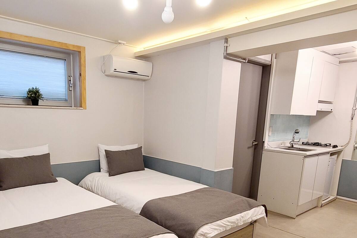 A compact bedroom with two beds, a small window with blinds, an air conditioning unit, and a kitchenette with a sink and cabinets.