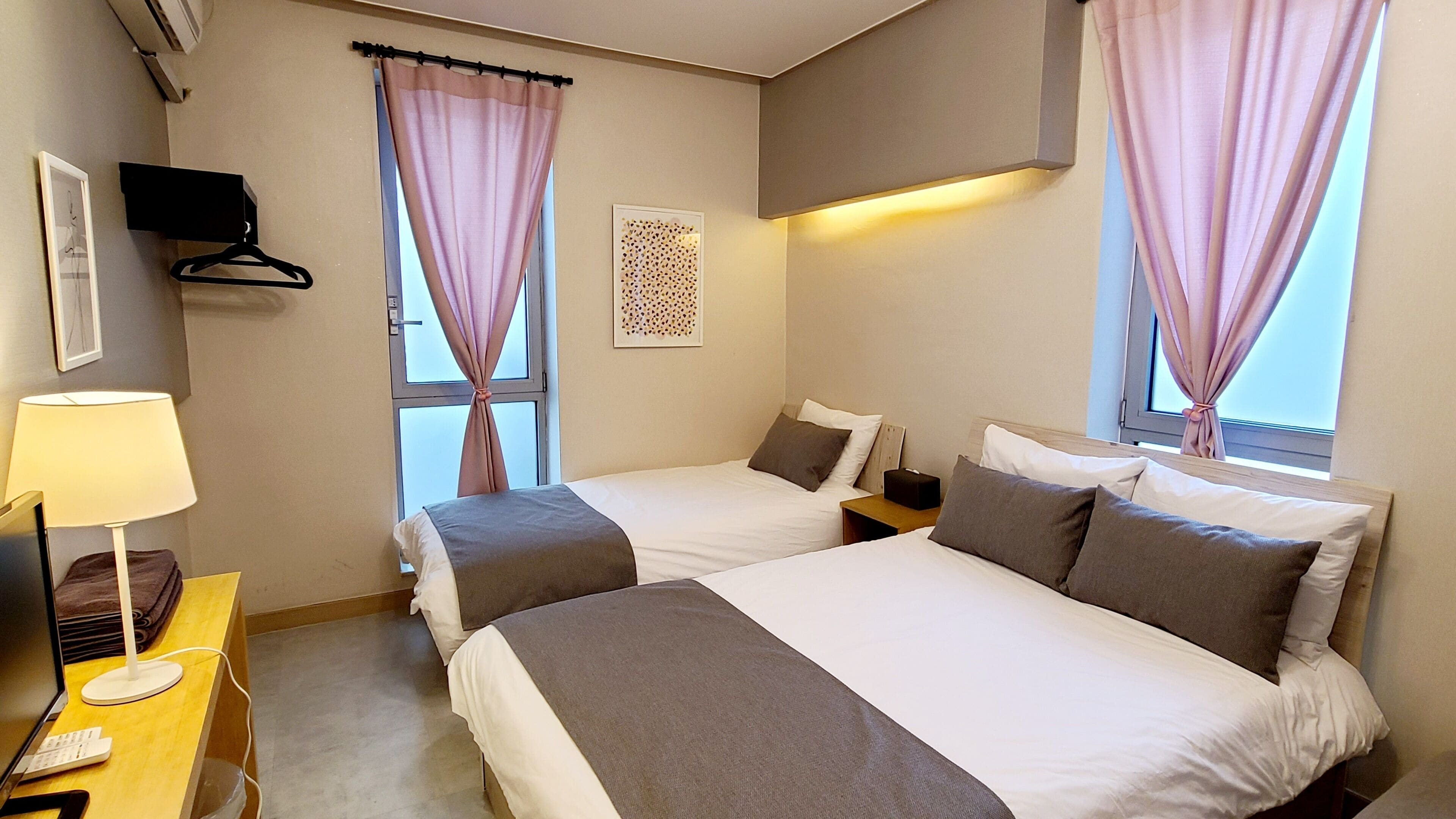 A hotel room with two beds, a desk with a lamp, a TV, and a window with curtains.