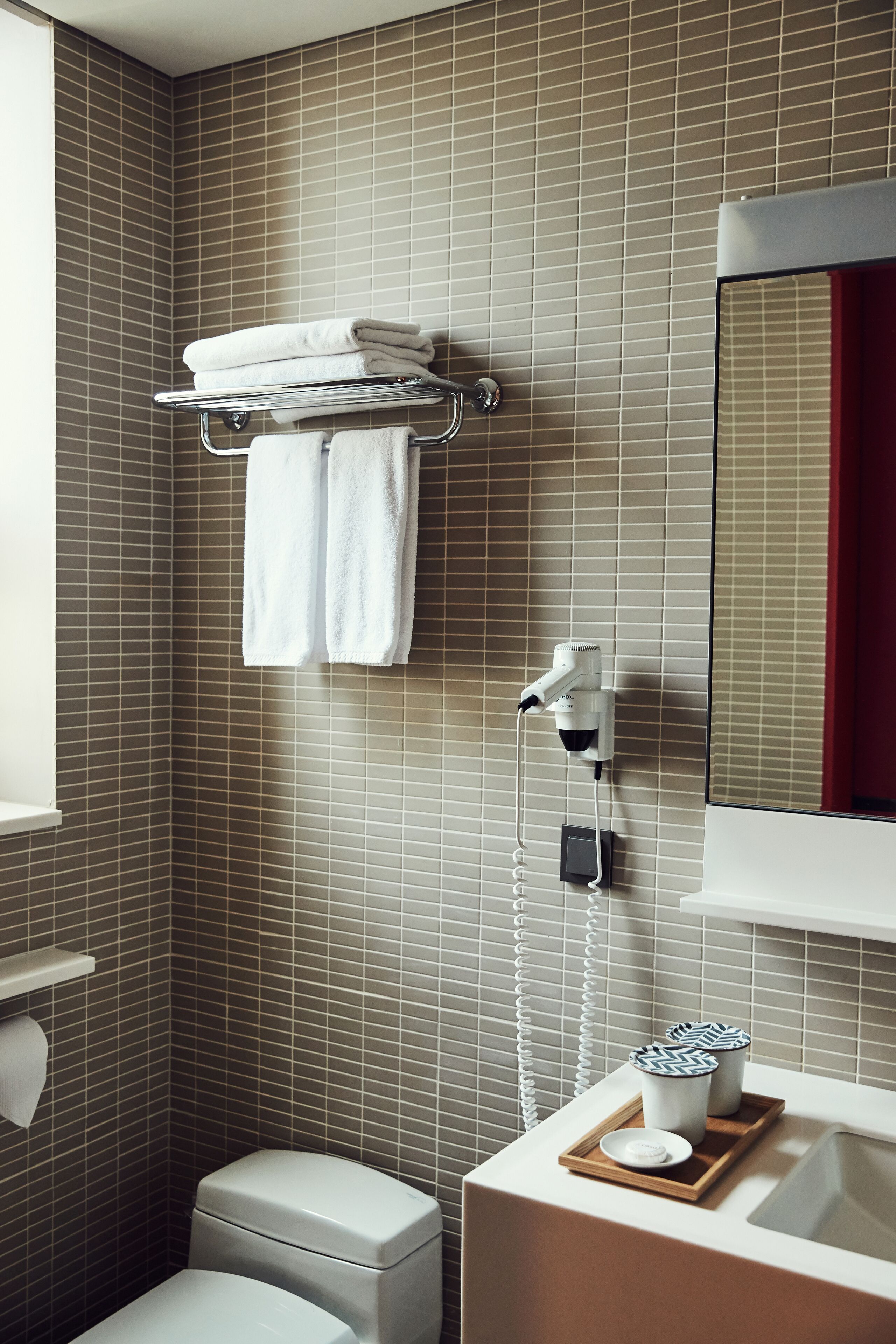 Un baño moderno con pared de azulejos, inodoro, lavamanos con bandeja y secador de pelo montado en la pared.