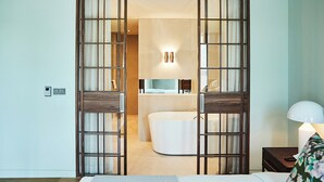 Habitación de hotel con una cama grande, una mesita de noche, una lámpara, un baño con bañera y un espejo.