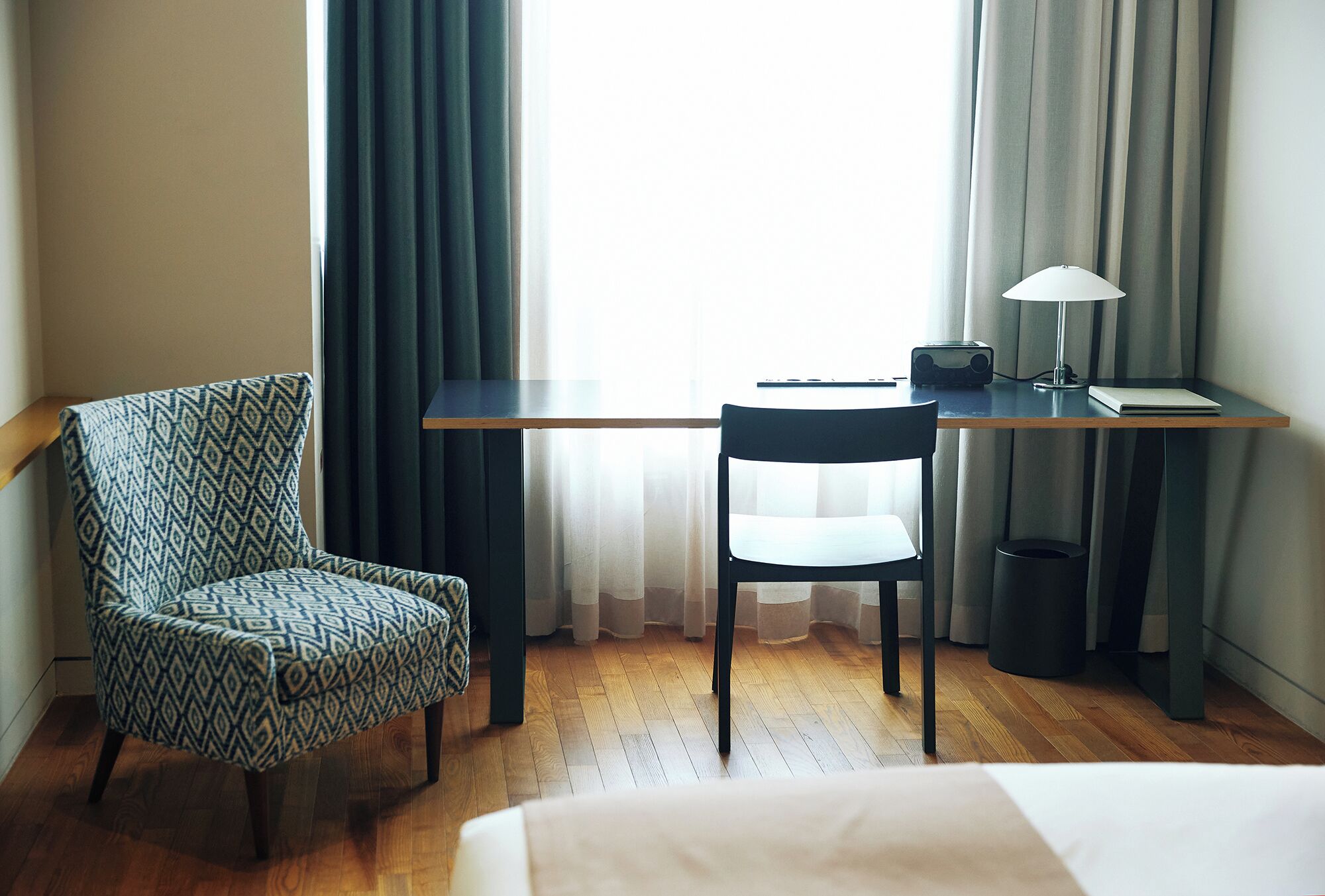 A hotel room with a bed, a desk with a lamp, a chair, and a window with curtains.