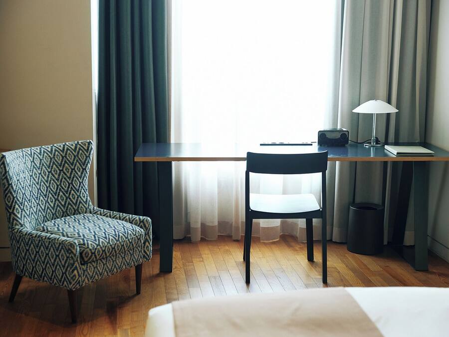 A hotel room with a bed, a desk with a lamp, a chair, and a window with curtains.