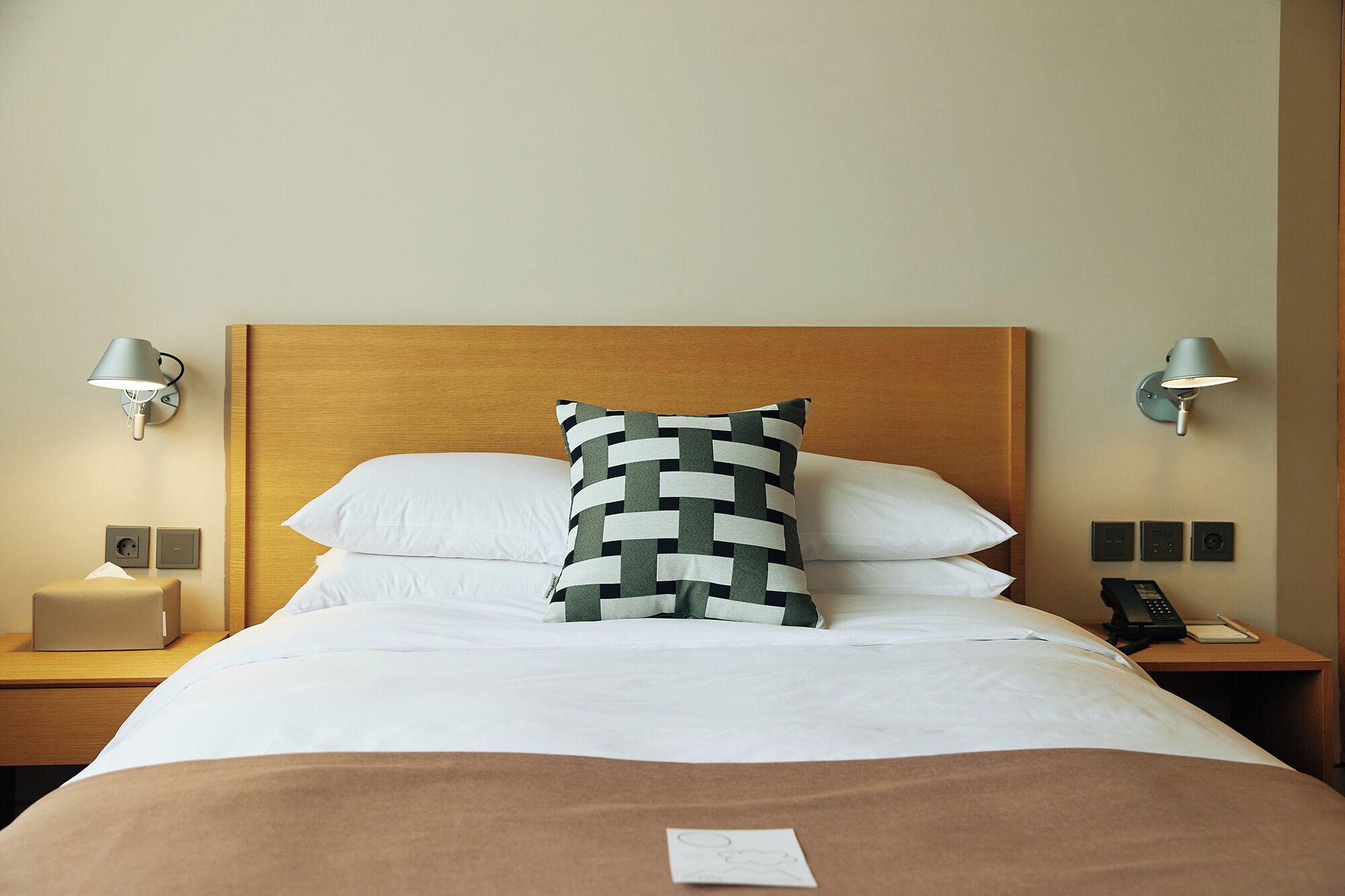 Ein Hotelzimmer mit einem hölzernen Kopfteil, weißer Bettwäsche, einem gemusterten Kopfkissen, Nachttischlampen, einem Nachttisch mit Lampe und einem Telefon.