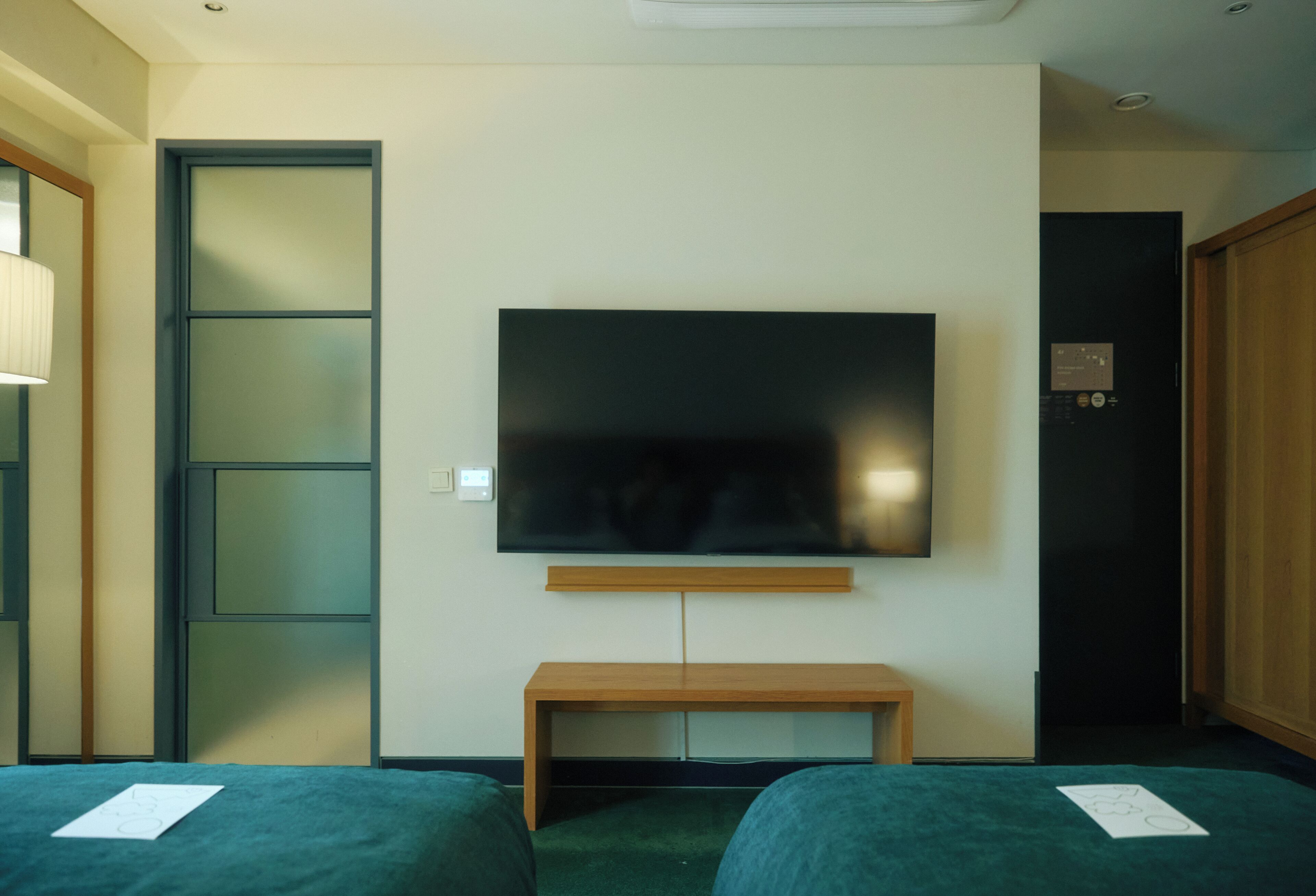 Une chambre d’hôtel dotée d’un téléviseur à écran plat fixé au mur, d’un banc en bois et de deux lits avec une literie verte.