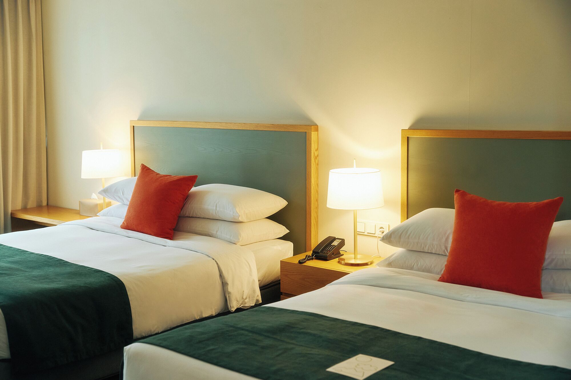 A hotel room with two beds, each with a green bedspread, white pillows, and red accent pillows. There are bedside lamps, a nightstand with a phone, and a wall-mounted lamp.