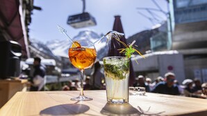 Uma mesa de madeira com duas bebidas, uma delas com a etiqueta 'MARTINI' e a outra com um ramo de hortelã, num ambiente ao ar livre com uma montanha e um teleférico ao fundo.