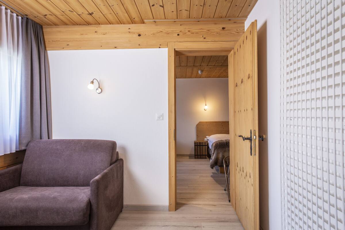 A room with a wooden ceiling, a bed with a headboard, a chair, and a wall-mounted light fixture.