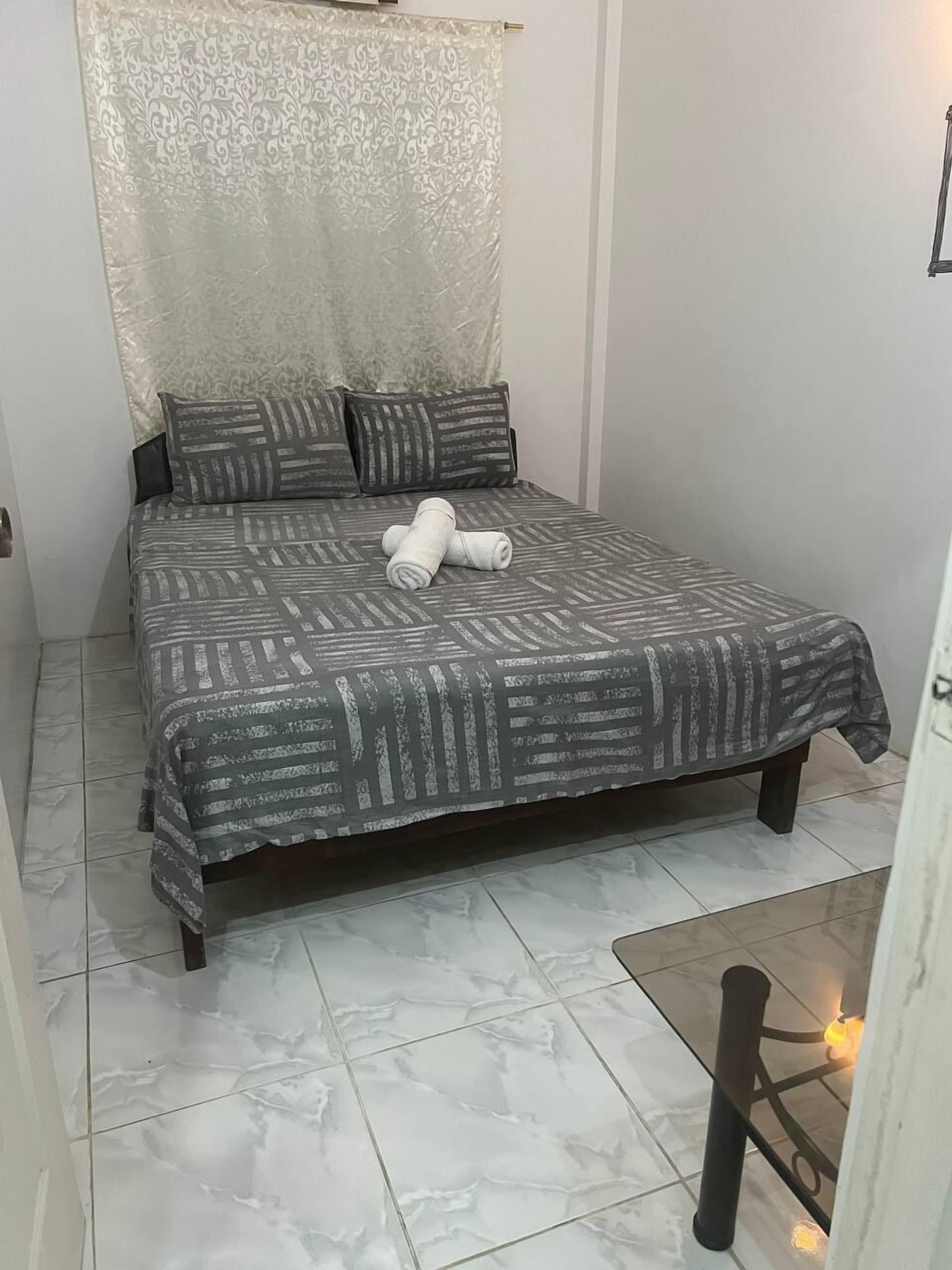 A small, neatly made bed with a patterned bedspread, a pair of rolled towels, and a decorative curtain.