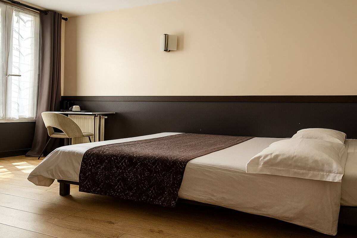 A hotel room with a bed, a chair, a small table, and a wall-mounted light fixture.