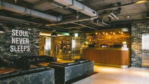 A modern hotel lobby with a stone wall, a reception desk, leather seating, and a sign that reads "SEOUL NEVER SLEEPS".