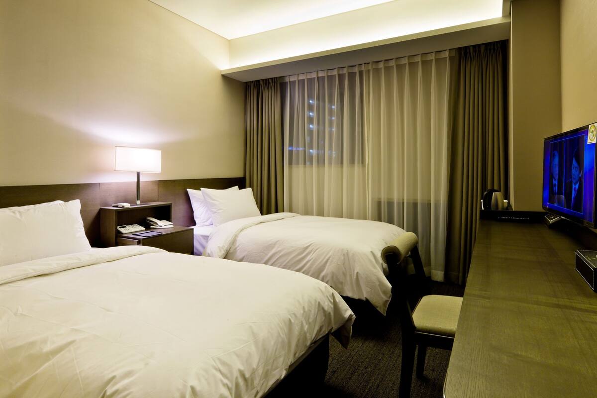 A hotel room with two beds, a desk with a television, a nightstand with a lamp, and a window with curtains.