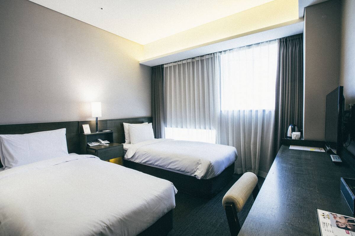A hotel room with two beds, a desk, a chair, a TV, and a window with curtains.