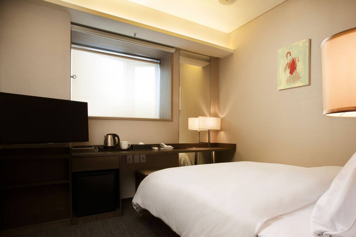 A hotel room with a bed, a desk with a flat-screen TV, a coffee maker, a lamp, and a painting on the wall.
