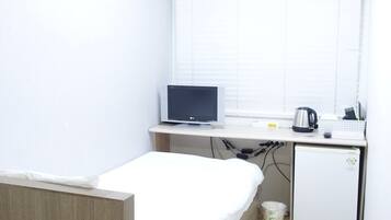 Una habitación pequeña y limpia con una cama individual, un escritorio con computadora, un refrigerador pequeño y una ventana con persianas.