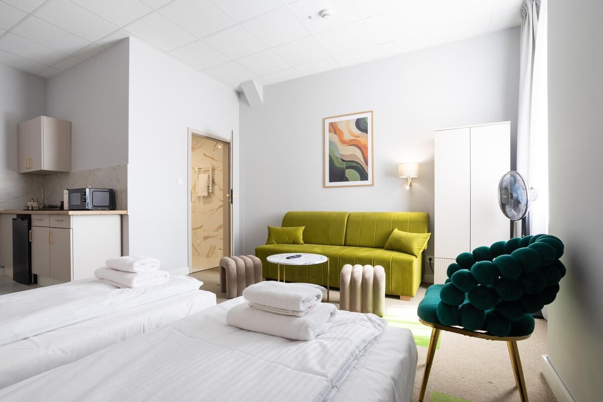 A hotel room with two beds, a green sofa, a round coffee table, a green chair, a painting on the wall, and a desk with a computer.