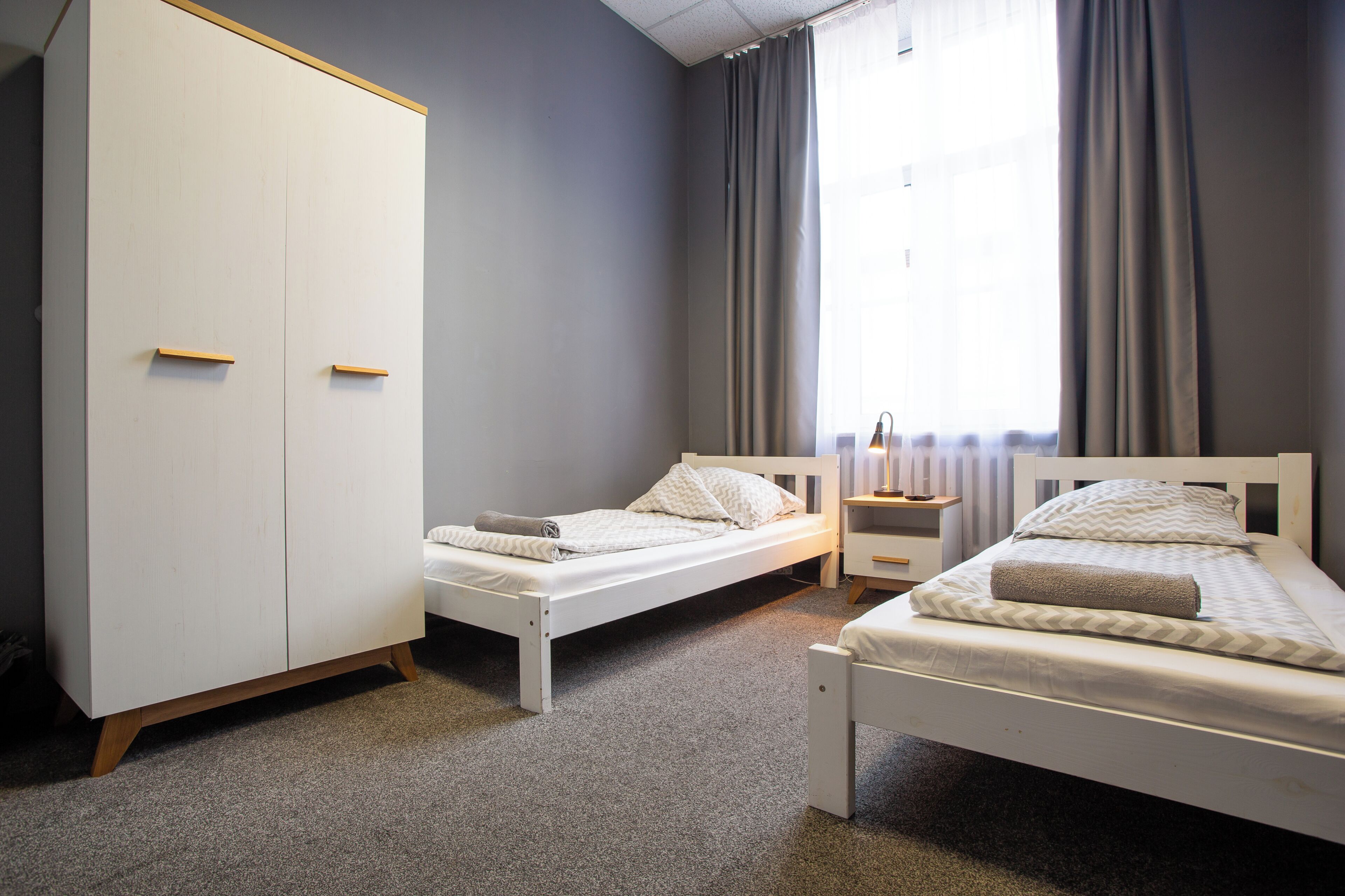 Une chambre avec deux lits simples, une armoire, une table de chevet et une fenêtre avec des rideaux.