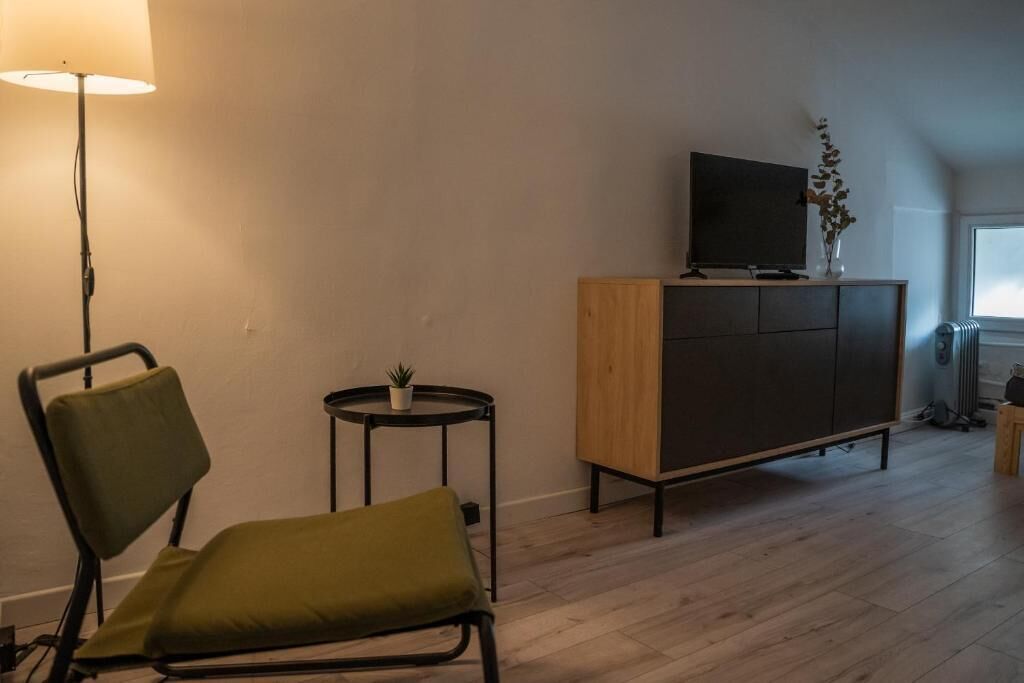 A room with a wooden cabinet, a television on top, a small table with a plant, and a chair with a cushion.