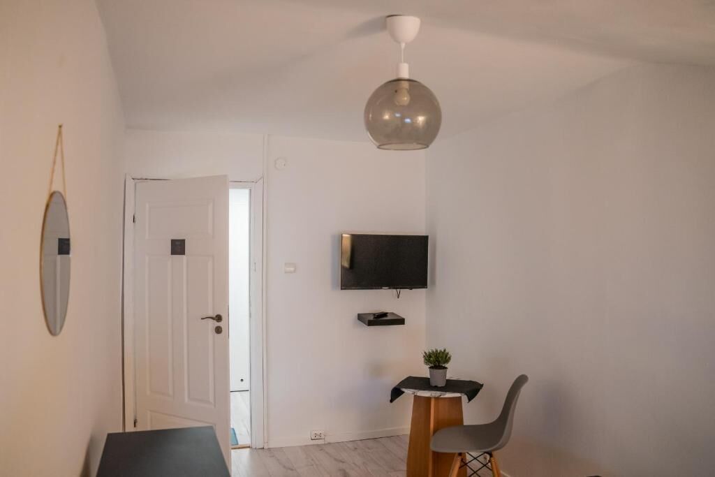 Una habitación moderna con una puerta blanca, un televisor montado, una pequeña mesa con una planta y una silla gris.