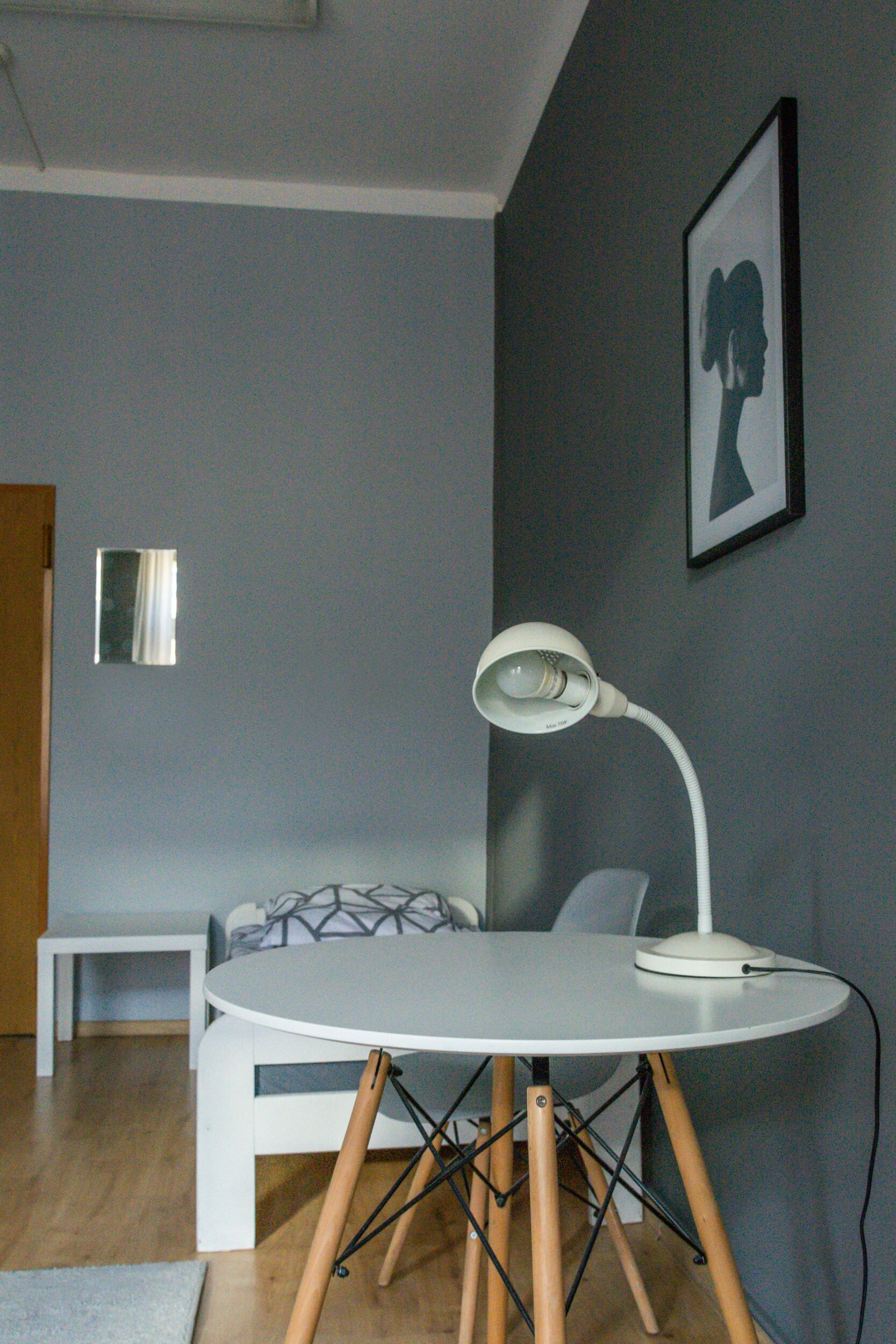 Una habitación moderna con una mesa redonda blanca, un escritorio blanco, una cama con ropa de cama estampada, una obra de arte enmarcada en la pared y un espejo.