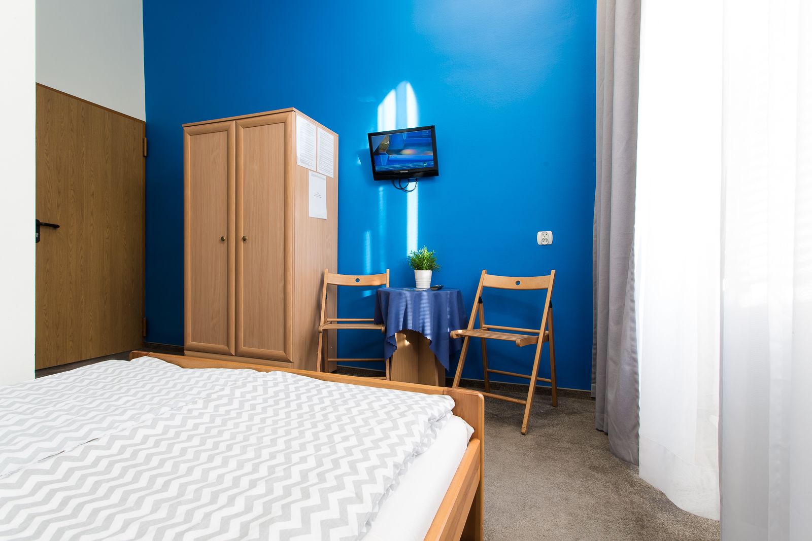 A bedroom with a bed, wooden wardrobe, a TV mounted on the wall, and a small table with a chair.