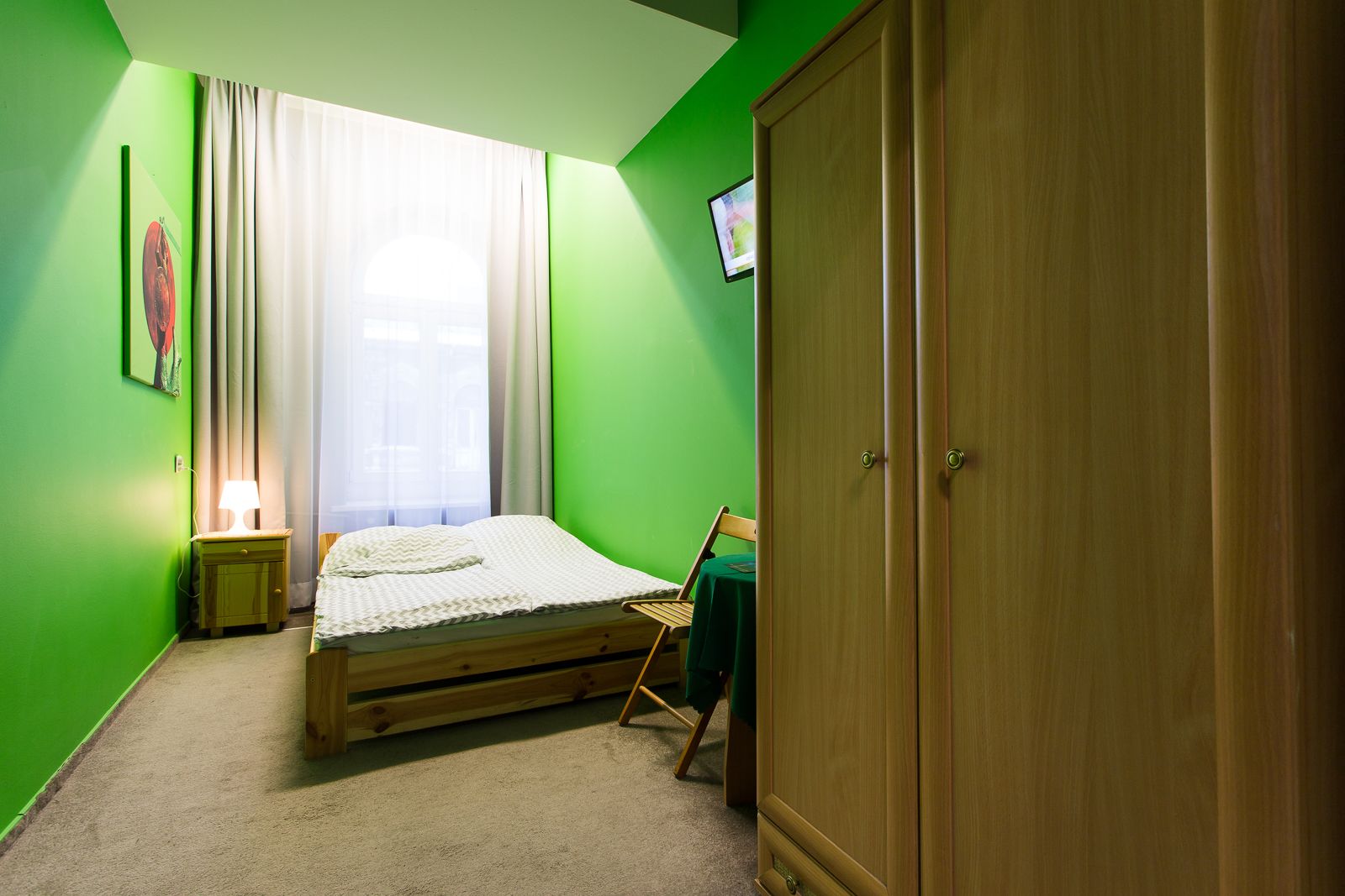 Una habitación con una cama, un armario de madera, un escritorio con una silla, una lámpara y un televisor en la pared.