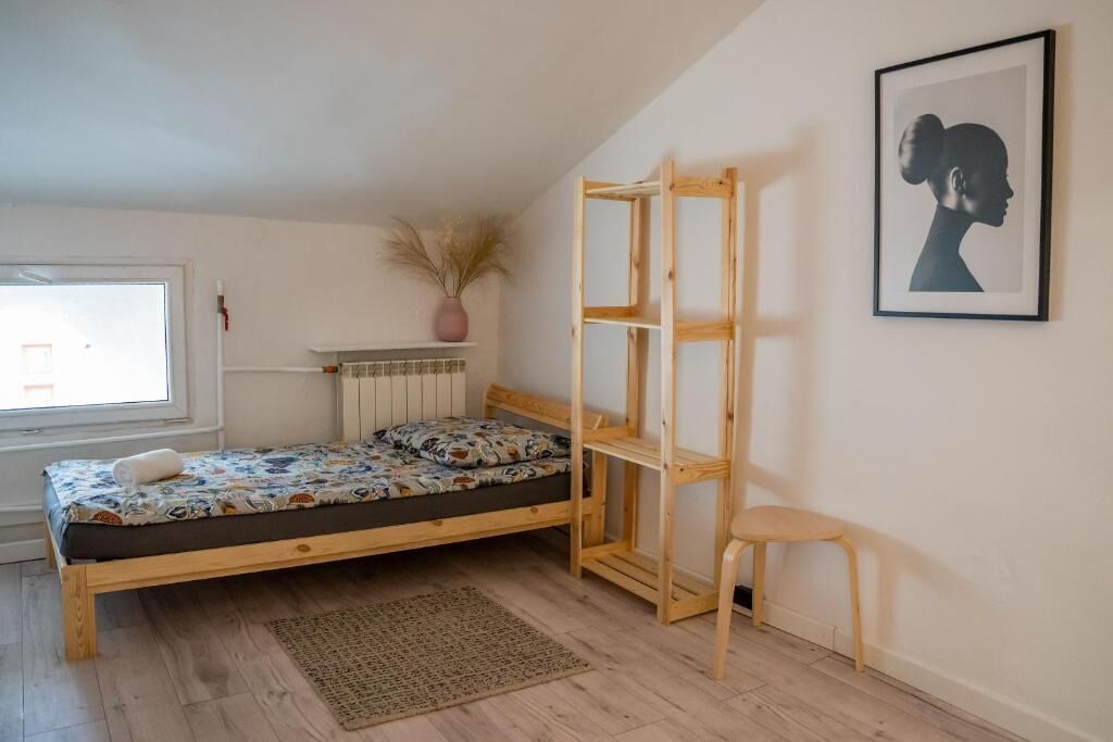 Une chambre avec un lit en bois, une petite chaise en bois, un radiateur, une fenêtre et un tableau encadré accroché au mur.