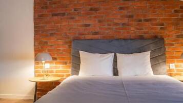 A bedroom with a sloped ceiling, exposed brick wall, a bed with white bedding, a grey headboard, and a small bedside table with a lamp.