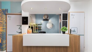 A reception desk with a wooden finish, a white counter, and a blue wall with a circular mirror. There are shelves with decorative items and a potted plant on the counter. - Moon Hostel Warszawa (Warsaw)