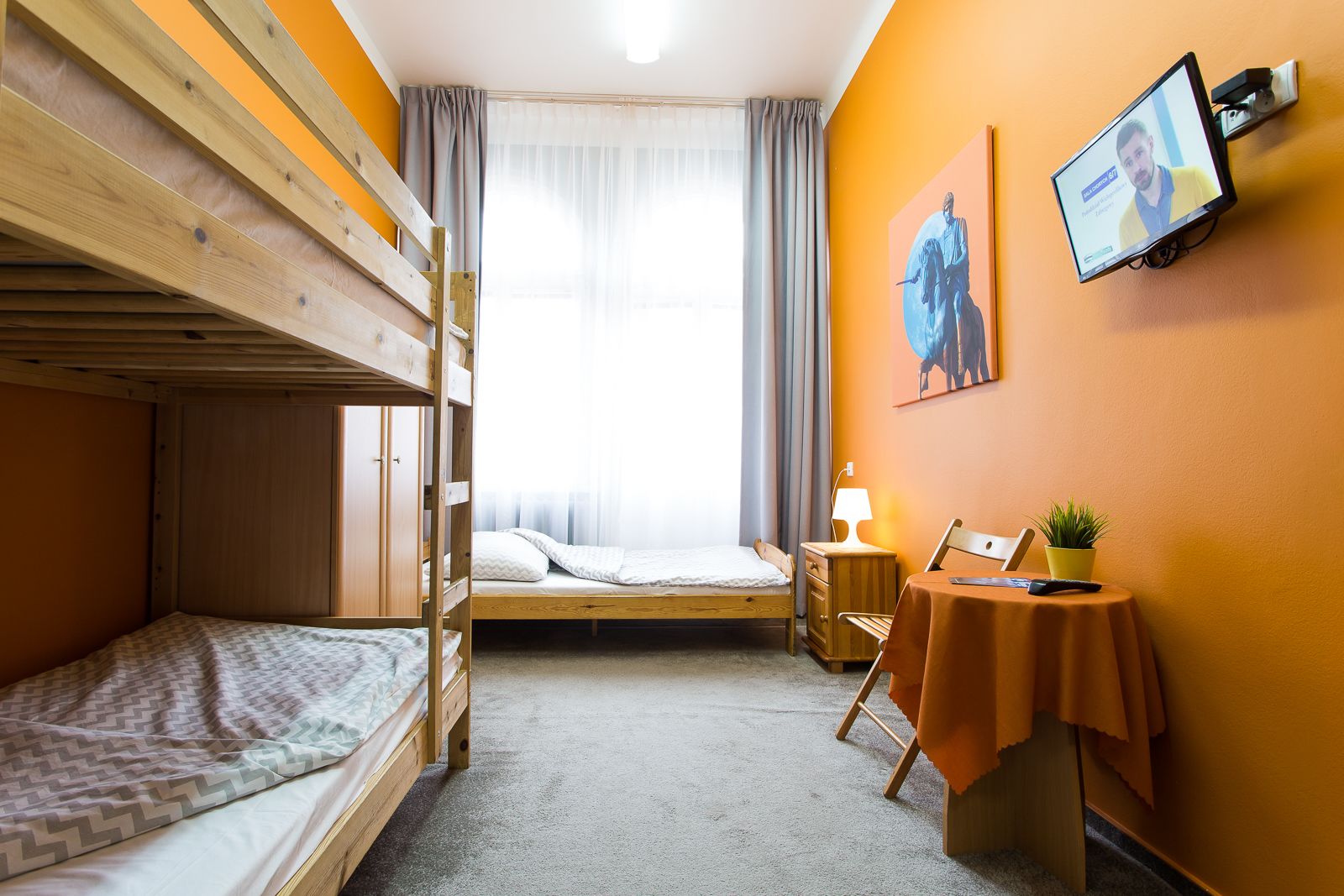 A room with bunk beds, a desk with a lamp, a small table with a plant, and a TV mounted on the wall.