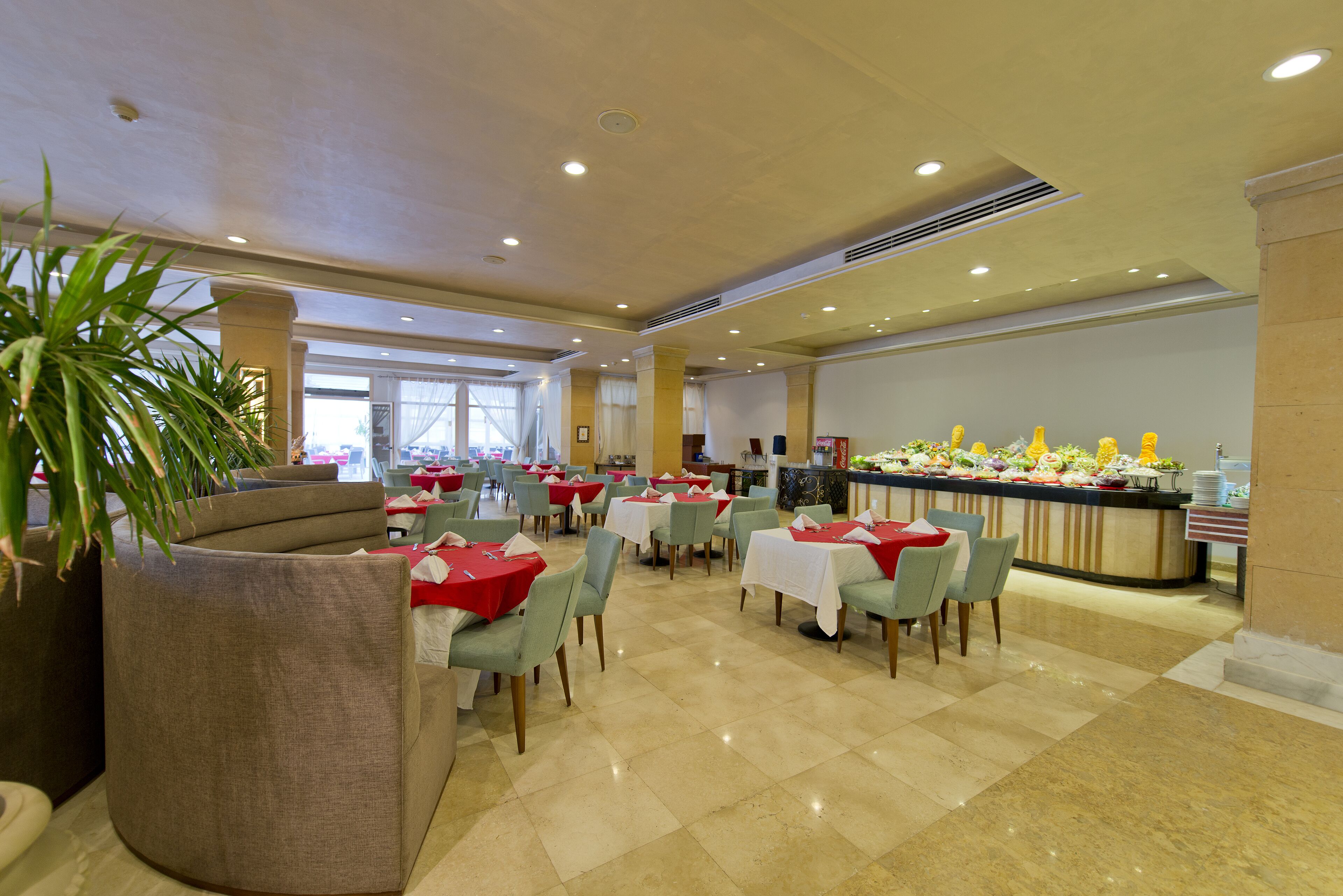 Un amplio comedor con múltiples mesas y sillas, un mostrador de buffet con diversos alimentos, y grandes macetas con plantas.