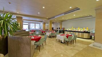Un amplio comedor con múltiples mesas y sillas, un mostrador de buffet con diversos alimentos, y grandes macetas con plantas.