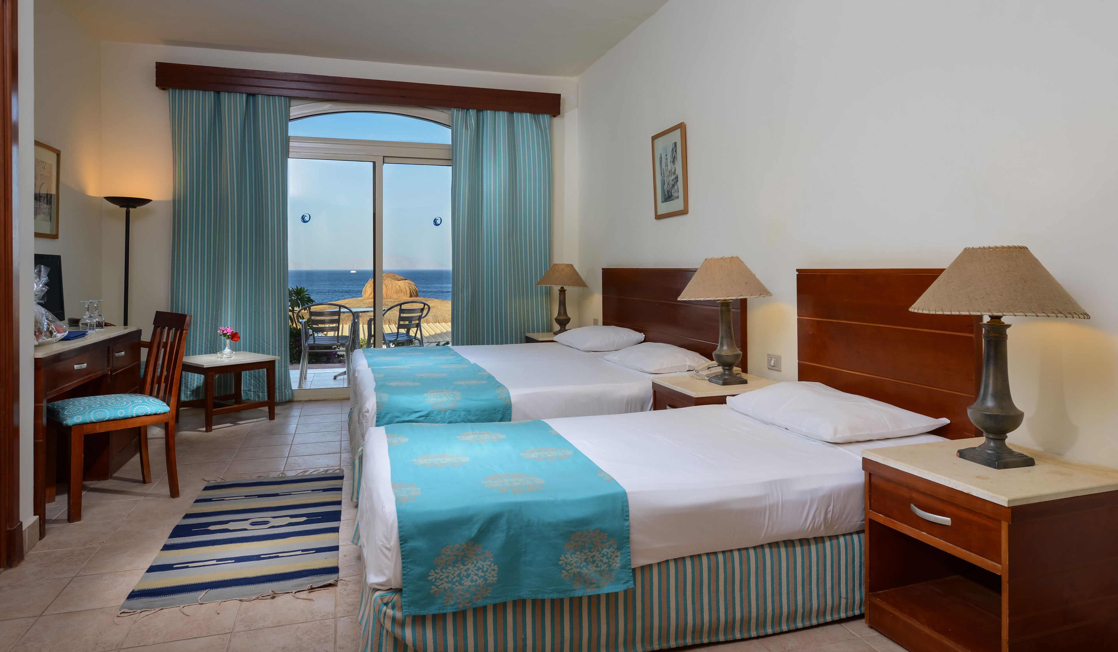 Habitación de hotel con dos camas, un escritorio, una silla, una lámpara y vista al mar.