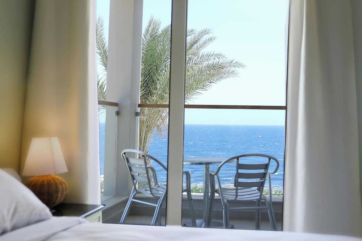 A room with a bed, a nightstand with a lamp, and a view of the ocean through large windows.