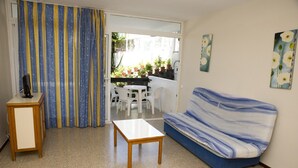 A living room with a sofa, a table, and a TV on a stand. There is a balcony with potted plants and a dining area with chairs. - Roca Verde (San Bartolomé de Tirajana)